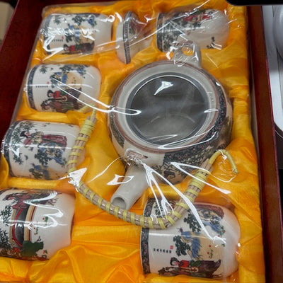 Set of Asian ceramic teapot and cups with decorative designs in clear plastic packaging on a yellow cloth.